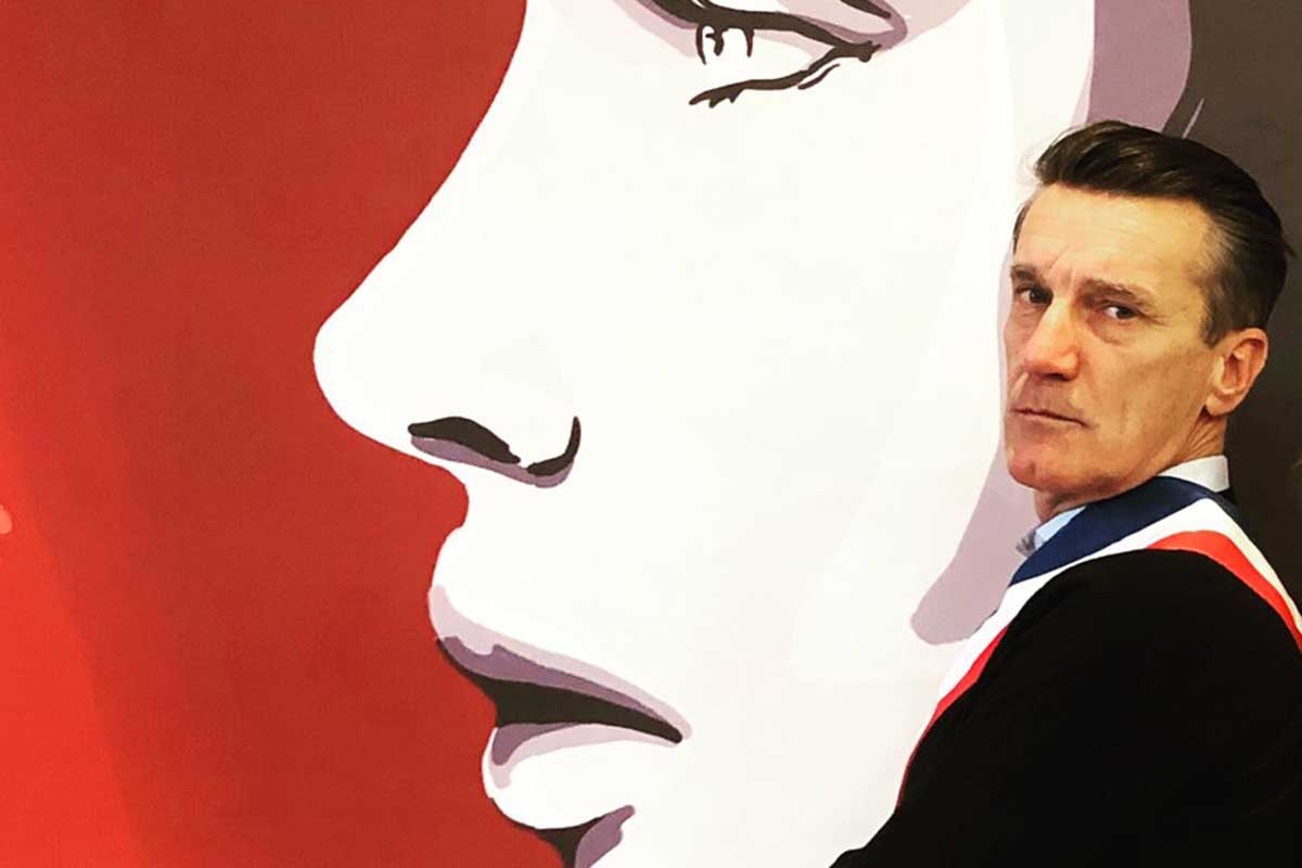 Bruno Ficheux pose de profil, avec son écharpe de maire. Derrière, on voit une reproduction du visage de Marianne qui symbolise la République française.